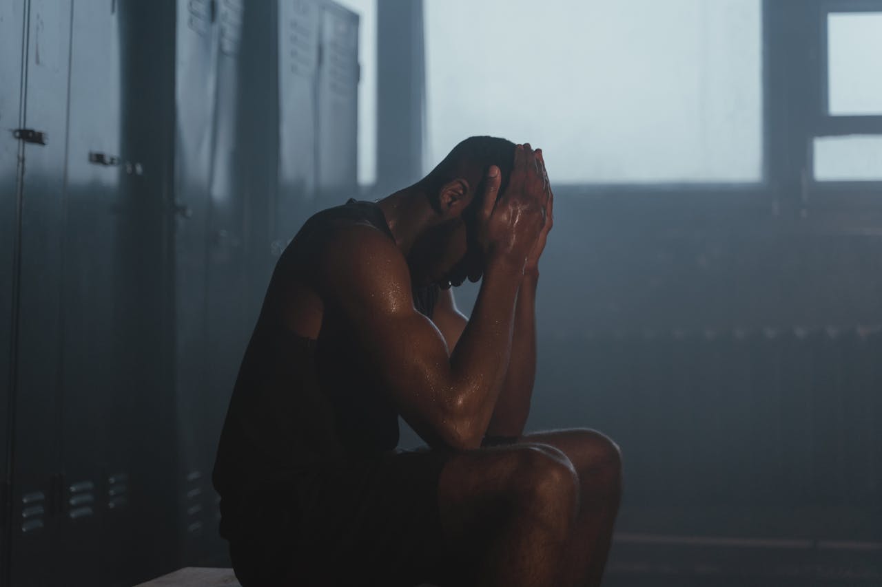 Sweaty man resting on bench in dimly lit locker room, showing exhaustion and recovery.